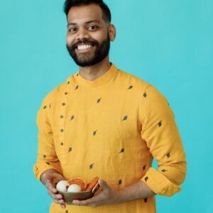 A cheerful man wearing a yellow kurta holding a bowl of traditional Indian sweets against a blue background.