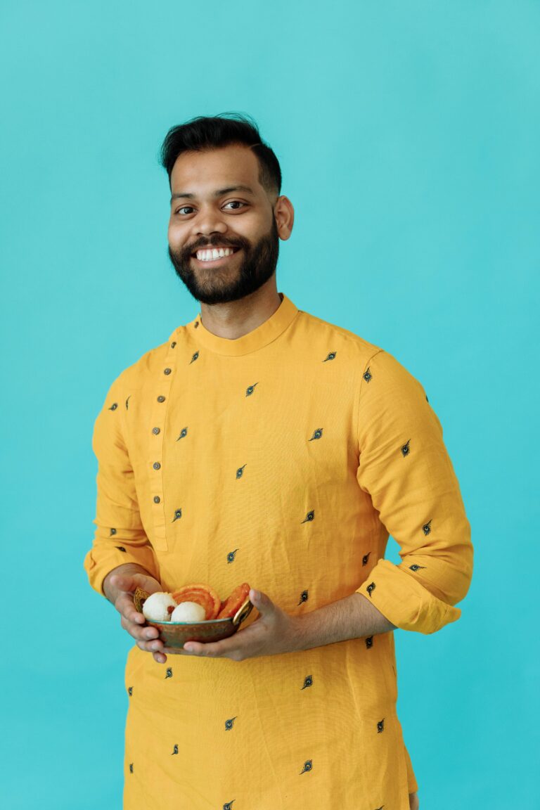 A cheerful man wearing a yellow kurta holding a bowl of traditional Indian sweets against a blue background.
