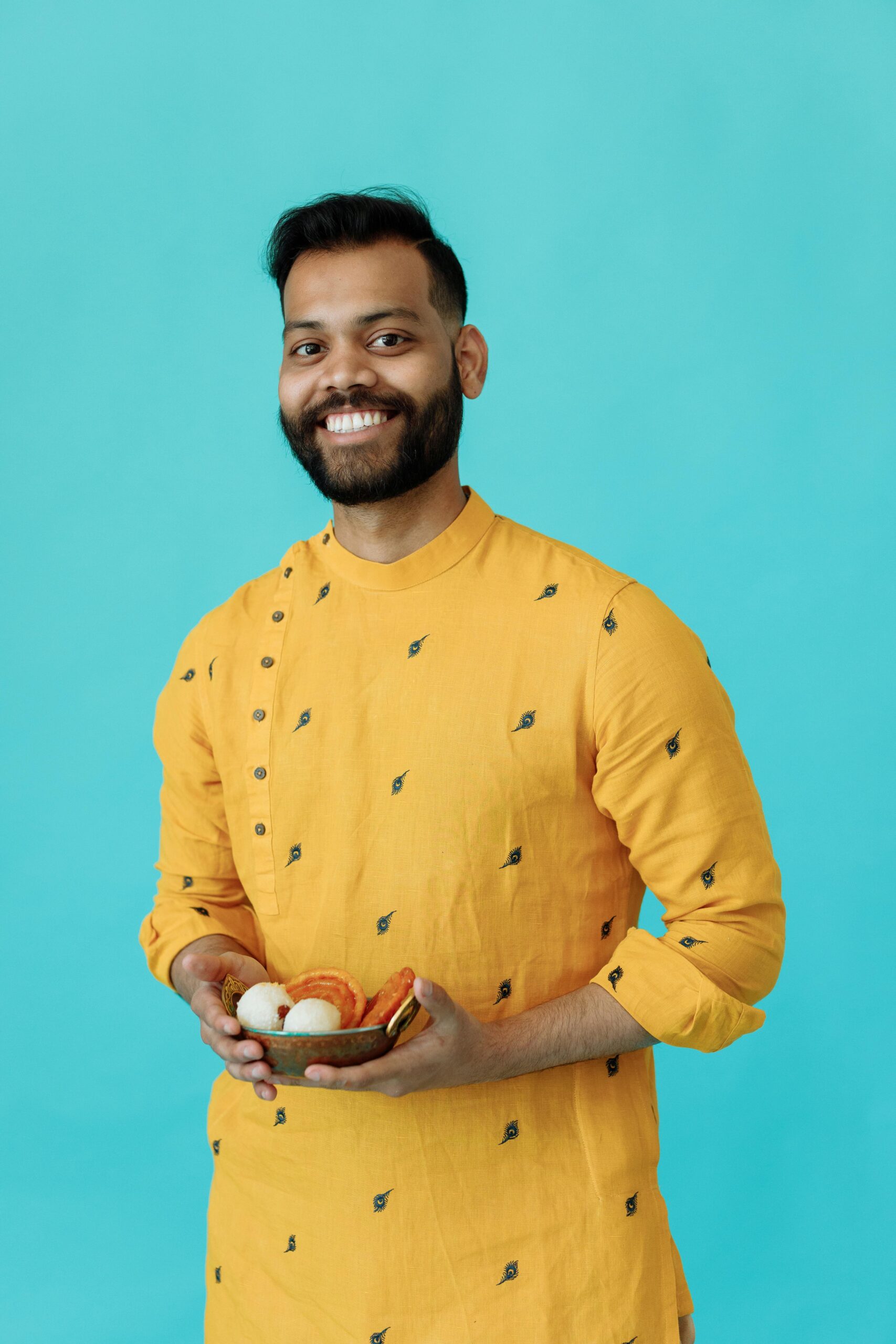 pexels photo 8489776 8489776 A cheerful man wearing a yellow kurta holding a bowl of traditional Indian sweets against a blue background.